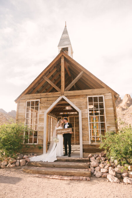 bride and groom in front of wooden chapel in the desert kissing nd holding just married sign