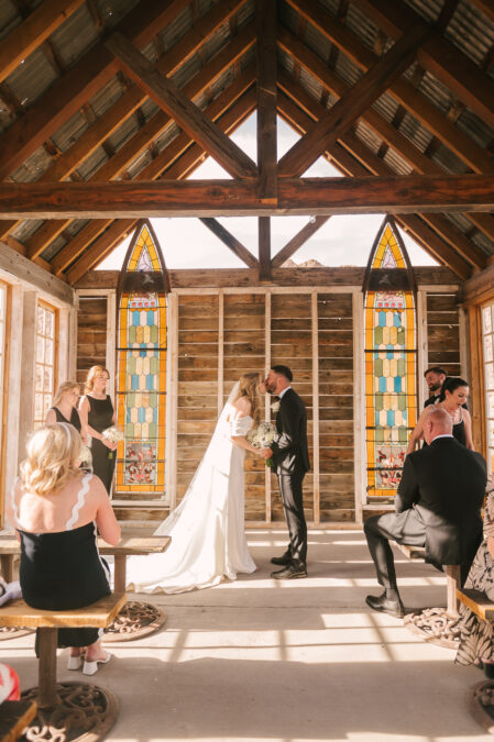 bride and groom first kiss las vegas desert wedding ceremony in wooden chapel with stained glass windows